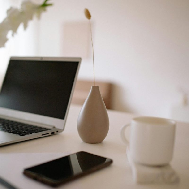 A clean and modern workspace featuring a laptop, vase, cup, smartphone, and paper on a white table.